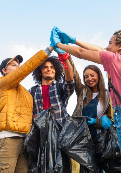 The image shows a group of enthusiastic volunteers celebrating their successful environmental cleanup. The team, consisting of a diverse mix of individuals, is seen raising gloved hands together in a gesture of unity and accomplishment. They hold black trash bags, indicating the litter they have collected. The group includes a Caucasian man in a yellow jacket, an African American man with curly hair in a plaid shirt, an Asian woman in denim overalls, and a person with pinkish hair in a red top. Their faces are filled with smiles, reflecting pride in their positive impact on the environment. Volunteer Group Celebrating Cleanup Success. High quality photo